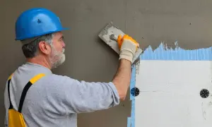 Pracownik w niebieskim kasku wykonuje gąbkowanie kleju na elewacji, przygotowując ją do dalszych prac.