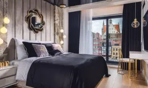 Sypialnia glamour inspiracje: luksusowe łóżko z czarną pościelą, aksamitne poduszki, ozdobne lustro i widok na zabytkowe kamienice.