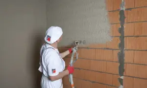 Pracownik w białym stroju nakłada tynk cementowo-wapienny na ścianę z pustaków ceramicznych za pomocą agregatu.