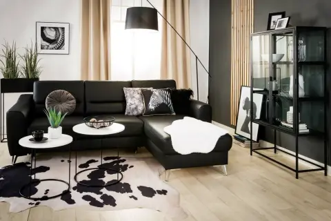 Nowoczesny salon z czarną ścianą w tle. Czarna skórzana kanapa, białe stoliki kawowe, dywan w cętki i szklana witryna tworzą elegancką przestrzeń.
