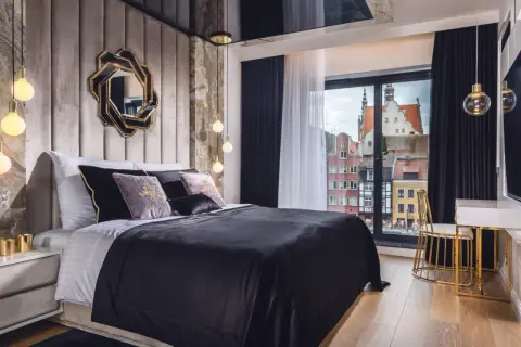 Sypialnia glamour inspiracje: luksusowe łóżko z czarną pościelą, aksamitne poduszki, ozdobne lustro i widok na zabytkowe kamienice.