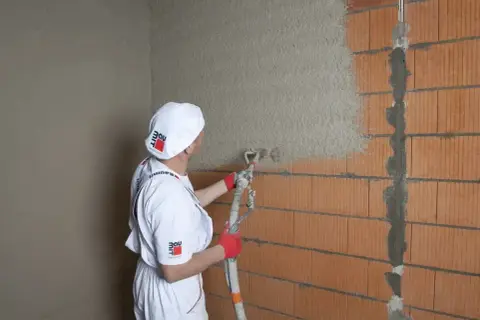 Pracownik w białym stroju nakłada tynk cementowo-wapienny na ścianę z pustaków ceramicznych za pomocą agregatu.