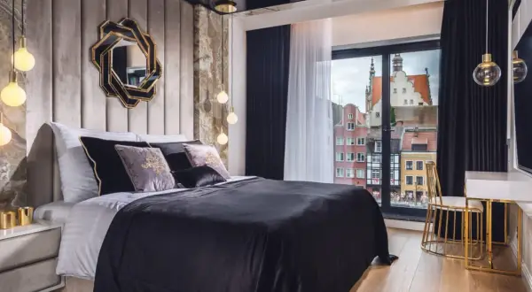Sypialnia glamour inspiracje: luksusowe łóżko z czarną pościelą, aksamitne poduszki, ozdobne lustro i widok na zabytkowe kamienice.