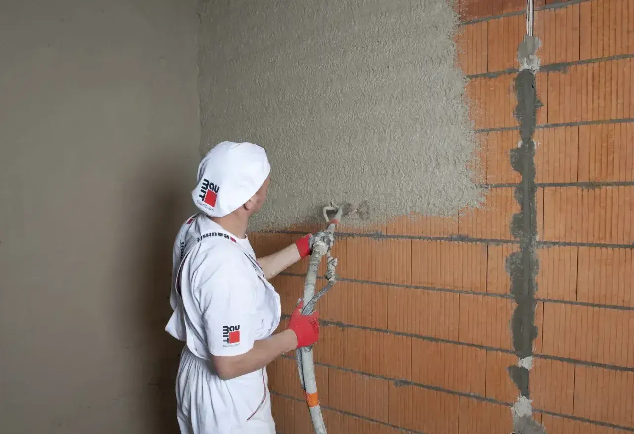 Pracownik w białym stroju nakłada tynk cementowo-wapienny na ścianę z pustaków ceramicznych za pomocą agregatu.