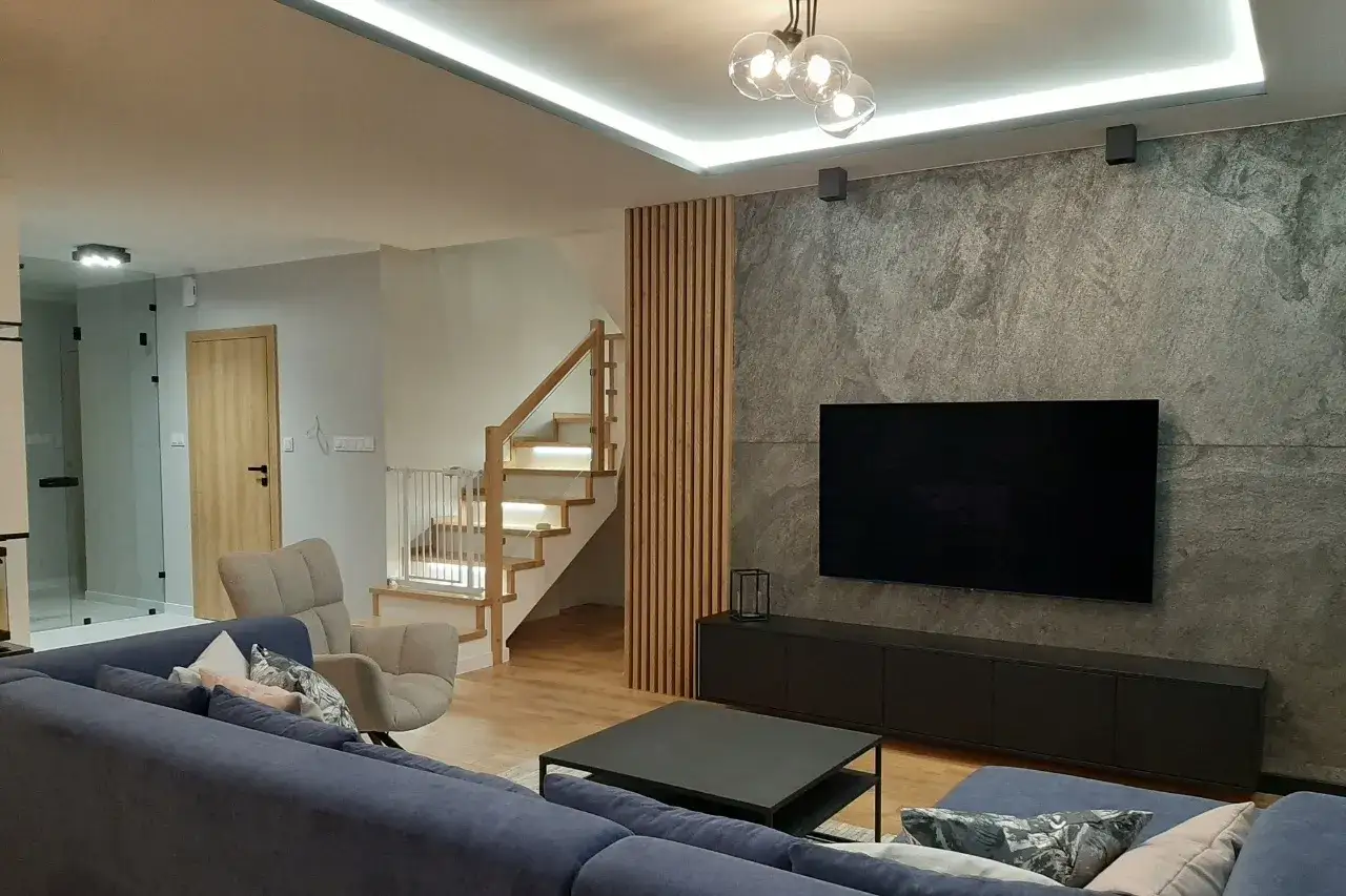 Nowoczesny salon z szarą kamienną ścianą i drewnianymi lamelami. Kanapa, fotel, schody i stolik kawowy tworzą przytulną przestrzeń. Lamele ścienne inspiracje.