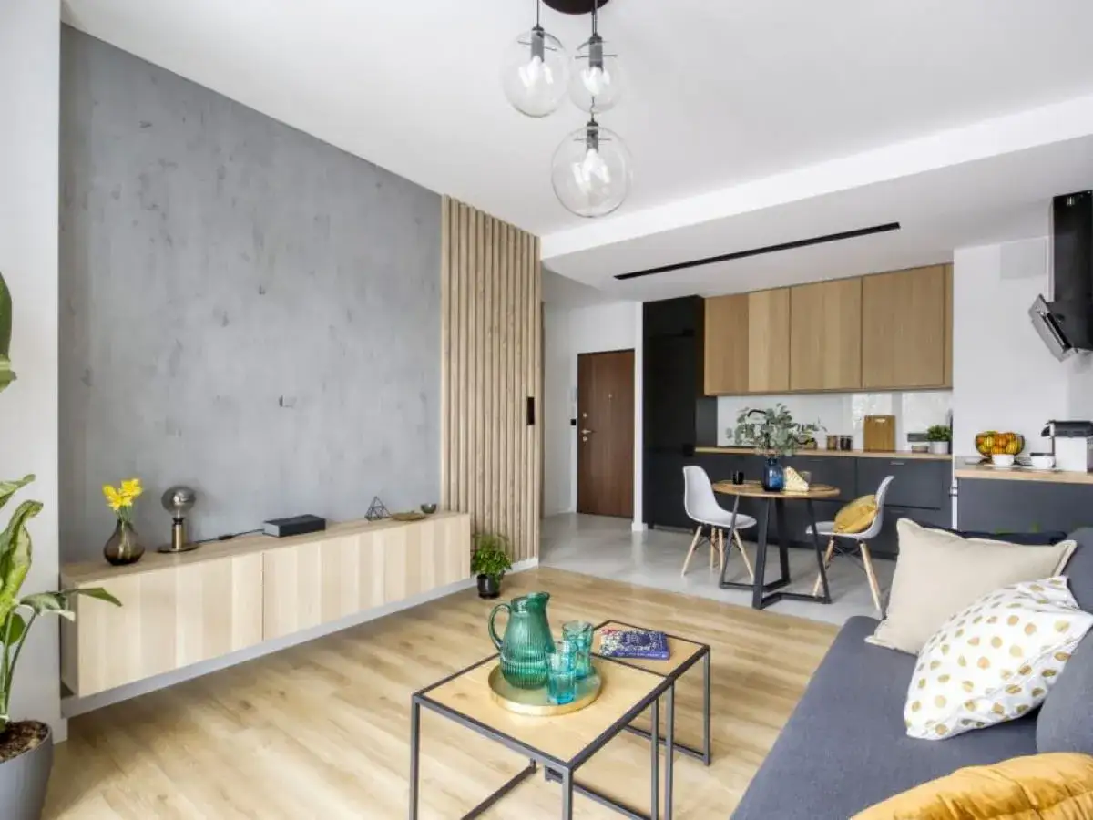 Nowoczesny salon z aneksem kuchennym. Betonowa ściana i drewniane lamele tworzą przytulną atmosferę. Zobacz, z czym łączyć lamele, by stworzyć stylowe wnętrze.