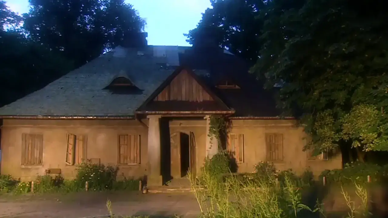 Stary dworek z rancza, otoczony drzewami, z uchylonymi okiennicami i otwartymi drzwiami.