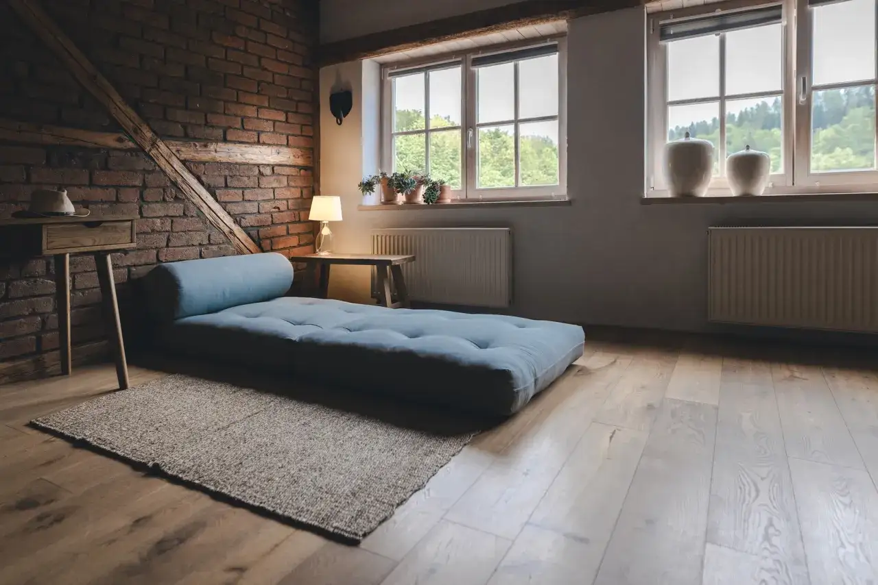 Niebieski futon na drewnianej podłodze, obok ceglanej ściany i okien. Idealne miejsce do odpoczynku, zastanawiasz się futon co to?