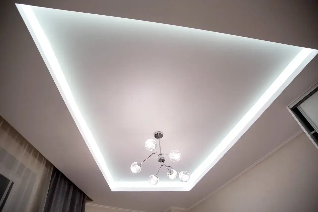 Nowoczesny sufit napinany wzory z podświetleniem LED tworzy trójkątny kształt. Centralnie wisi designerska lampa.