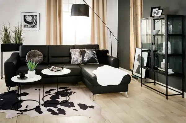 Nowoczesny salon z czarną ścianą w tle. Czarna skórzana kanapa, białe stoliki kawowe, dywan w cętki i szklana witryna tworzą elegancką przestrzeń.